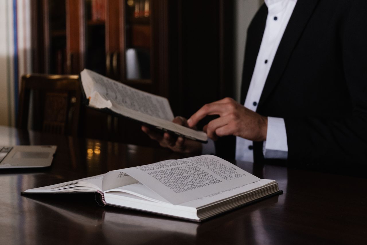 services-09 A man in a suit reading religious books at a wooden table indoors.